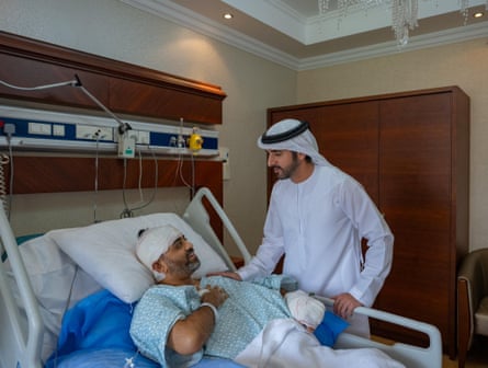 The crown prince, wearing a white robe and headdress, stands over the hospital bed talking to the pilot, whose head and left arm and hand are heavily bandaged.