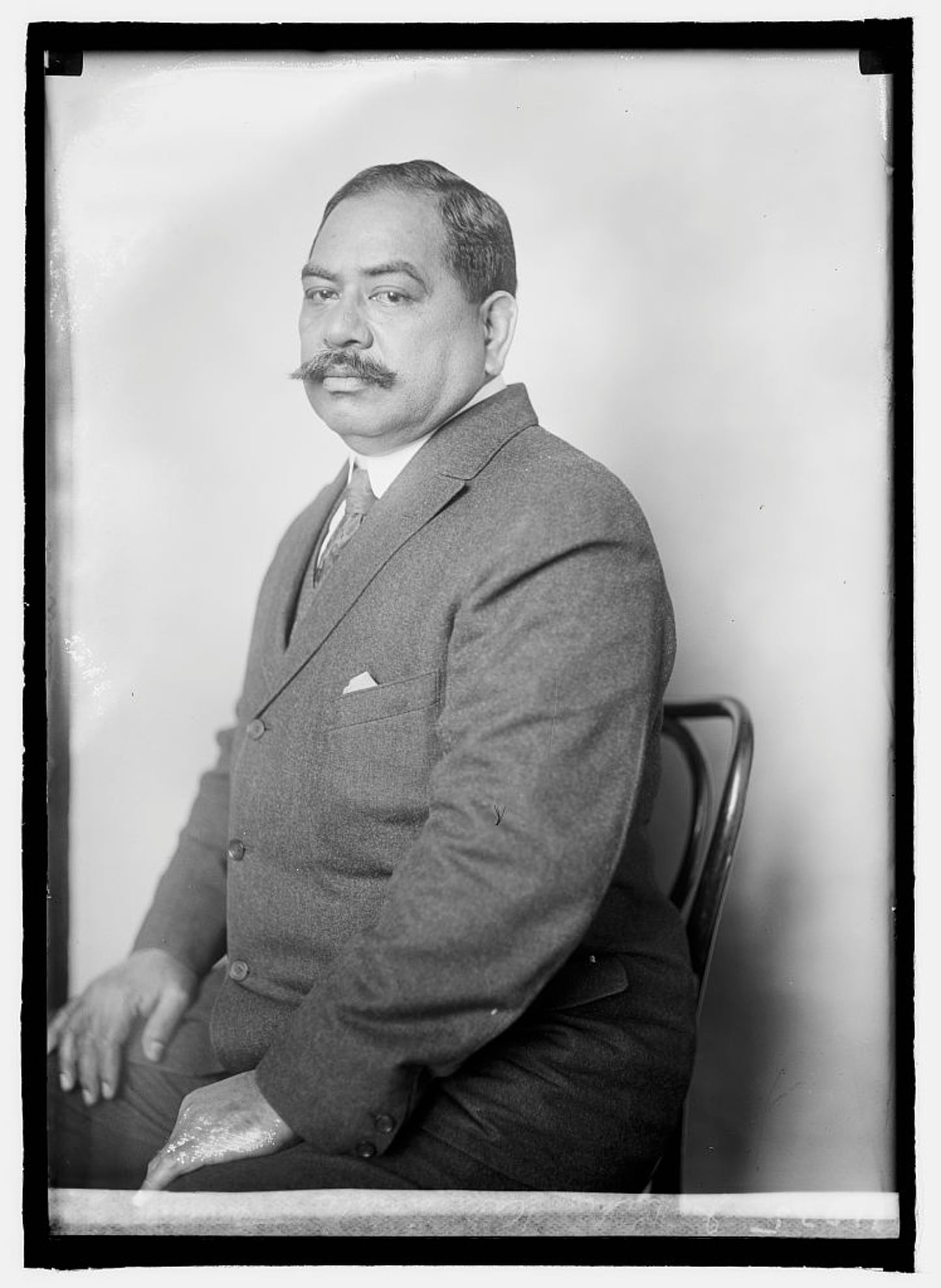 Black-and-white photo of a Native Hawaiian man with short dark hair, a twirly tipped moustache, and a suit, sitting in a cane-backed wooden chair.
