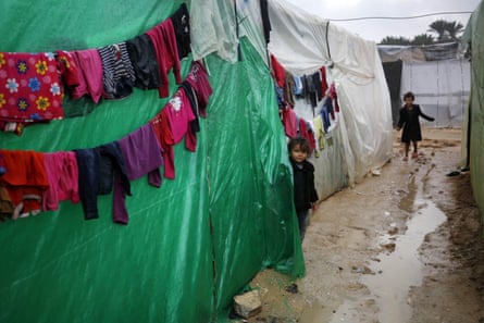 Two small children look at the camera in an muddy alleyway with a big puddle between shelters made from sheets of plastic. A line of clothing is hanging up to dry.