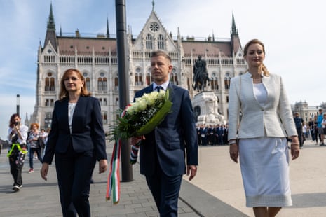 Candidate for parliament Agnes Forsthoffer (right), incoming prime minister Péter Magyar (centre) and and Andrea Bujdosó, leader of the Tisza faction in parliament (left) arrive at Parliament in Budapest, walking in a row with Magyar holding a wreath of white flowers. Behind them is a blue sky and a building.