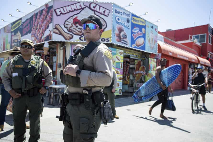 Sheriff's deputies in full police gear and weapons stand next to a funnel cake shack on Venice boardwalk.