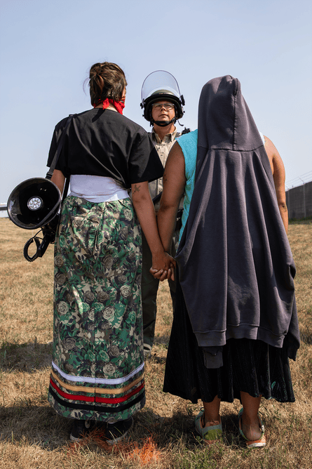 The backs of two people, one in indigenous wear, who are standing in front of a policeman