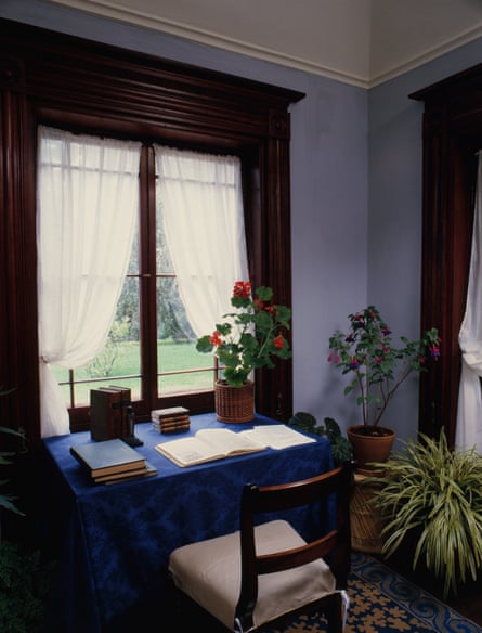 an old fashioned desk with an open book and pot of geraniums on it, in front of a window