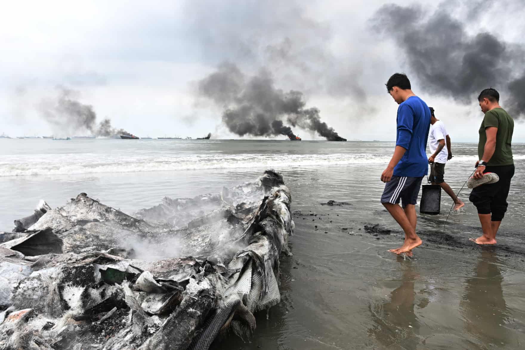 People walk on sand near water with smoke plumes in the background and a charred boat nearby