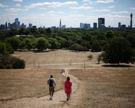 People walk across a path through dry, brown grass with trees and London's skyline in the distance