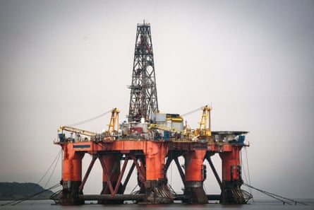 An anchored oil rig anchored in the Cromarty Firth. It is painted orange, with yellow cranes, and is seen against a dark grey sea and grey sky.