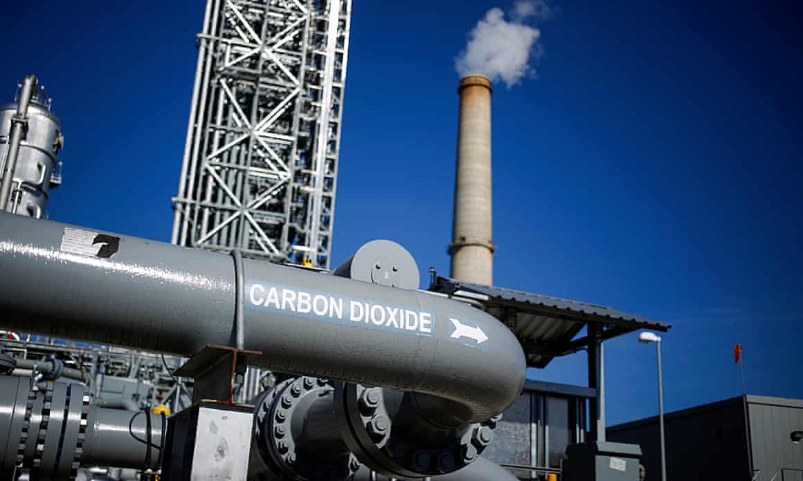 A large gray pipe labeled 'carbon dioxide' is photographed in front of a smoking smokestack and a tall white structure in Thompsons, Texas