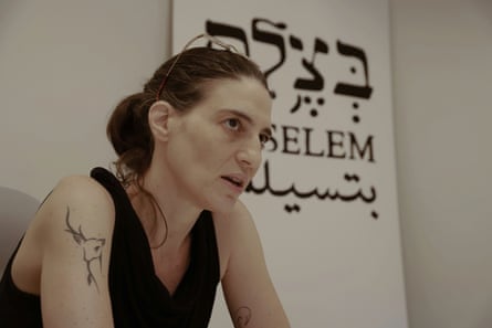 she has dark hair tied back in a ponytail and wears a sleeveless black top, showing a tattoo on her upper arm.