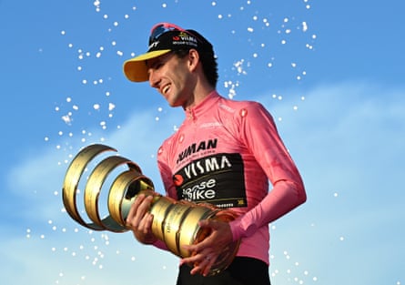 Simon Yates holds a big trophy which is a spiralling gold ribbon