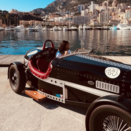 A woman sits in an old-fashioned open-top car by a waterfront with yachts and high-rise flats in the background