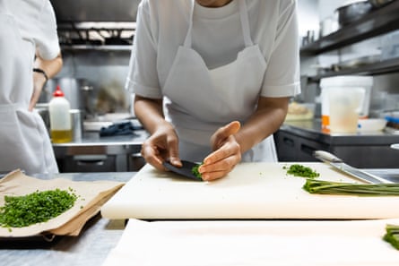 In a commercial kitchen, a chef wearing a white T-shirt and a white apron uses a bench scraper to pick up a pile of finely chopped chives off a cutting board