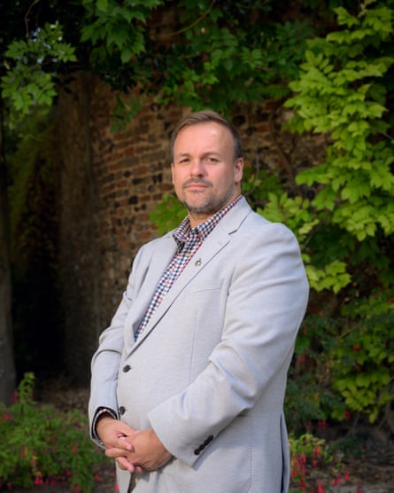 A man in his 40s with stuble and short dark hair stands for a portrait against a wall covered in shrubbery. He is wearing a light-grey suit and a checked shirt with no tie