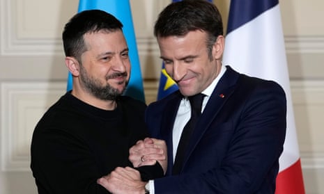 Volodymyr Zelenskiy and Emmanuel Macron shake hands while standing if front of a Ukrainian and French flag.