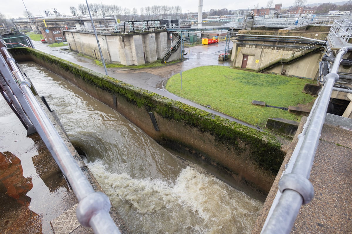 Brownish water gushes down a concrete channel at the sewage works, with small industrial buildings in the background.