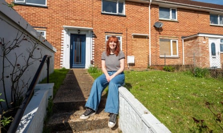 Laura nelson, in a T-shirt, jeans and trainers, sits on a concrete ledge next to grass in front of a brick building in the sun