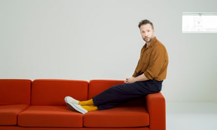 Rafe Spall studio shot, sitting on the arm of a red sofa, his feet on the seat