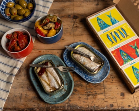 An image, taken from above, of a table with two tins of tinned fish, a bowl of green olives, a bowl of roasted red pepper and a bowl of roasted vegetables. There is also a box containing brightly coloured cards with pictures of fish on them.