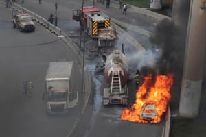 A car engulfed in flames on the side of a road. A fire engine and firefighter are nearby