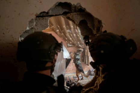 A dog sniffs curtains in a heavily damafged house as a soldier looks on through a large hole in the wall.