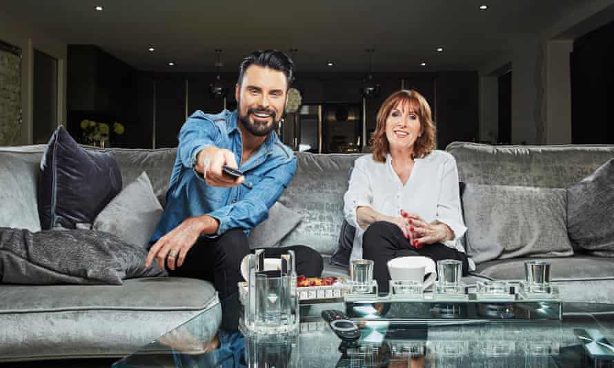 Rylan and his mother side by side on a large gray sofa, with Rylan pointing a Tv remote control straight at the camera