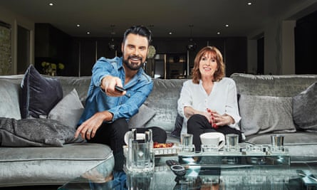 Rylan and his mother side by side on a large grey sofa, with Rylan pointing a Tv remote control straight at the camera