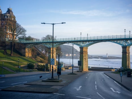 A footbridge of four braced iron arches on tapering stone piers over a road, with the sea in the distance and an old building on the left