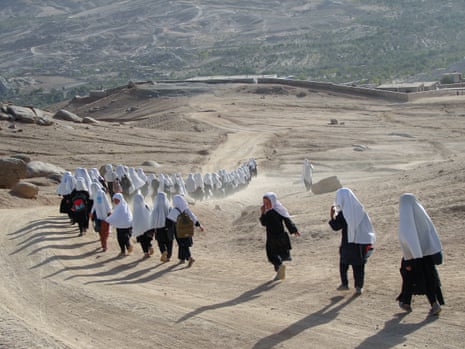 A line of girls in white hijabs walk down a dusty road