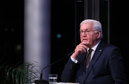 he is 70, with white hair and glasses, and wears a dark suit; he is illuminated in a dark room.