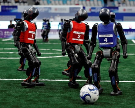 Robots in blue and red vests kick a ball about on an indoor pitch