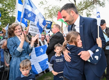 A man is hugged by a child as a woman and two other children look on, to the backdrop of Quebec flags.