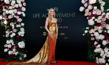 Nicole Kidman stands in front a board reading 'life achievement award' on the red carpet. She wears a sparkly gold dress that trails behind her.