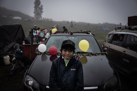 A boy attends the Tree Celebration festival in Queros. He is standing at the front of a car with balloons tied to the wipers, there is another car parked alongside and groups of people in the background. The sky is dark and the weather looks grim.