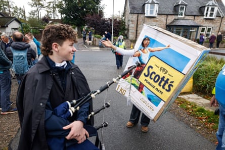 A woman wears a giant Scott’s Porage Oats cardboard box with a hole for her face. A young man carrying bagpipes looks on, smiling.
