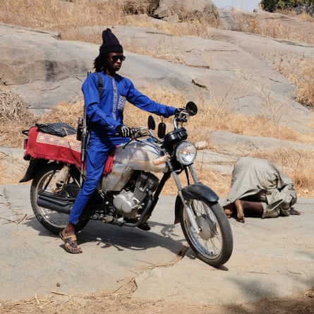 A gang member straddles his motorbike while a man prays next to him