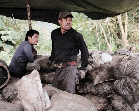Two men stand among piles of sacks beneath a tarpaulin