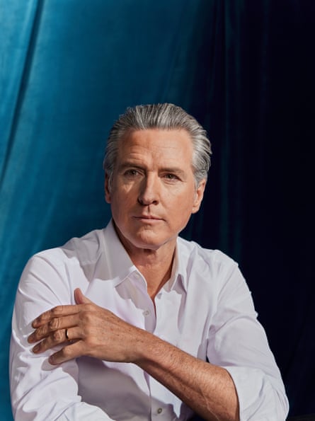 A shot of a seated Gavin Newsom from just above the waist up, front on to the camera, looking just to the right of it, with his left arm across his body, his left arm touching his right upper arm, wearing a white shirt with the sleeves rolled up and no tie, against a turquoise velvet background