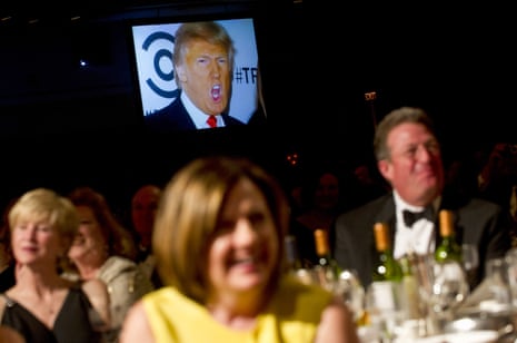 A man's face is displayed on a television screen above people in formal dress laughing as they sit at tables set with dinnerware and wine bottles.