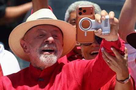 Lula da Silva, wearing a wide-brimmed hat, smiles while taking a selfie with his wife’s phone