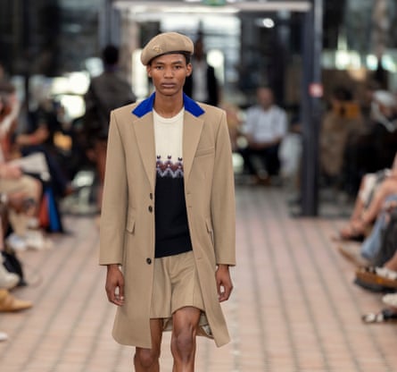 A model walking the catwalk wearing a patterned jumper, shorts, long structured coat and a beret.