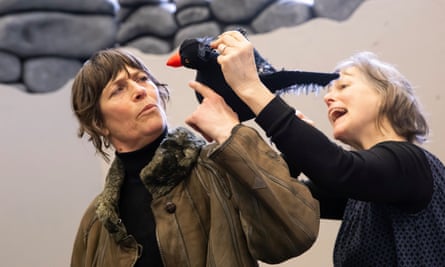 A woman talks to a puppet of a blackbird, puppeteered by another woman, in front of a faux stone wall