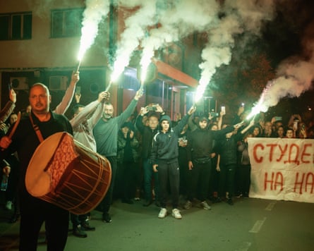 A welcome party of protesters raise flares and hold a banner and one beats a bass drum on the side of a road at night