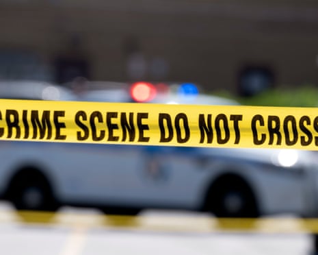 Yellow crime scene tape stretches across a parking area with a police car blurred behind it