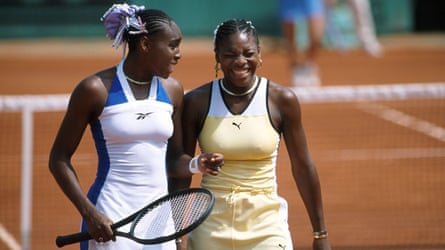 Venus dressed in tennis whites holding a racket and Serena Williams in white and yellow on court in front of the net.