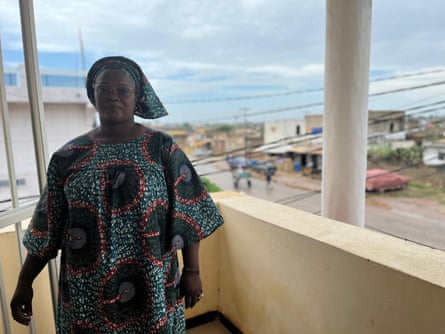 A middle-aged African woman in a traditional matching headscarf and dress, standing on a balcony with the sea in the distance.
