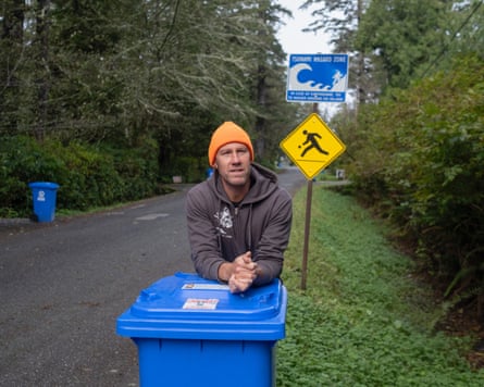 A man stands on a street resting his arms on a blue bin