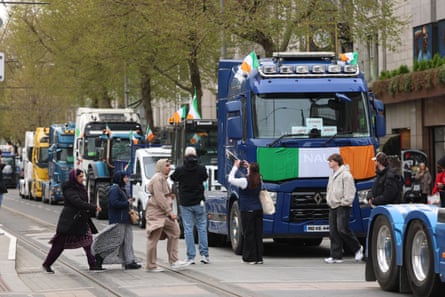 Trucks with Irish flags attached to them queue in a wide city street as people cross the road and take photographs.