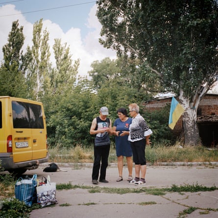 Three people stand on a street next to the back of a yellow van with bags of shopping on the ground near to it
