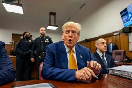 a man in a blue suit and yellow tie sits at a table with police officers behind him