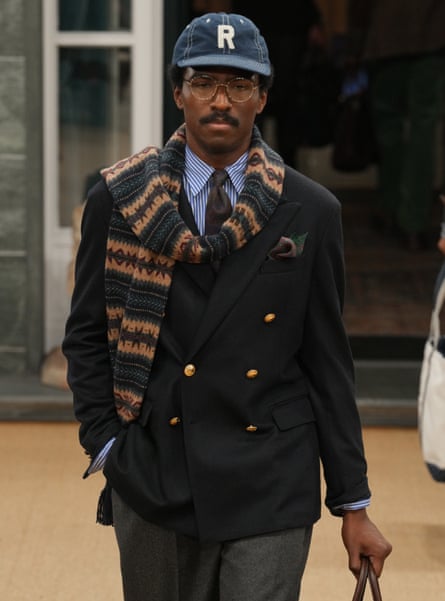 A model wears a shirt and tie under a double-breasted navy blazer, green-and-gold striped scarf and a denim cap branded with an R
