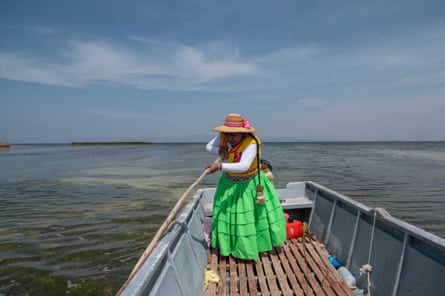 A woman in a bright green ankle-length dress and a straw hat pushes her boat through the shallow water with a wooden pole.
